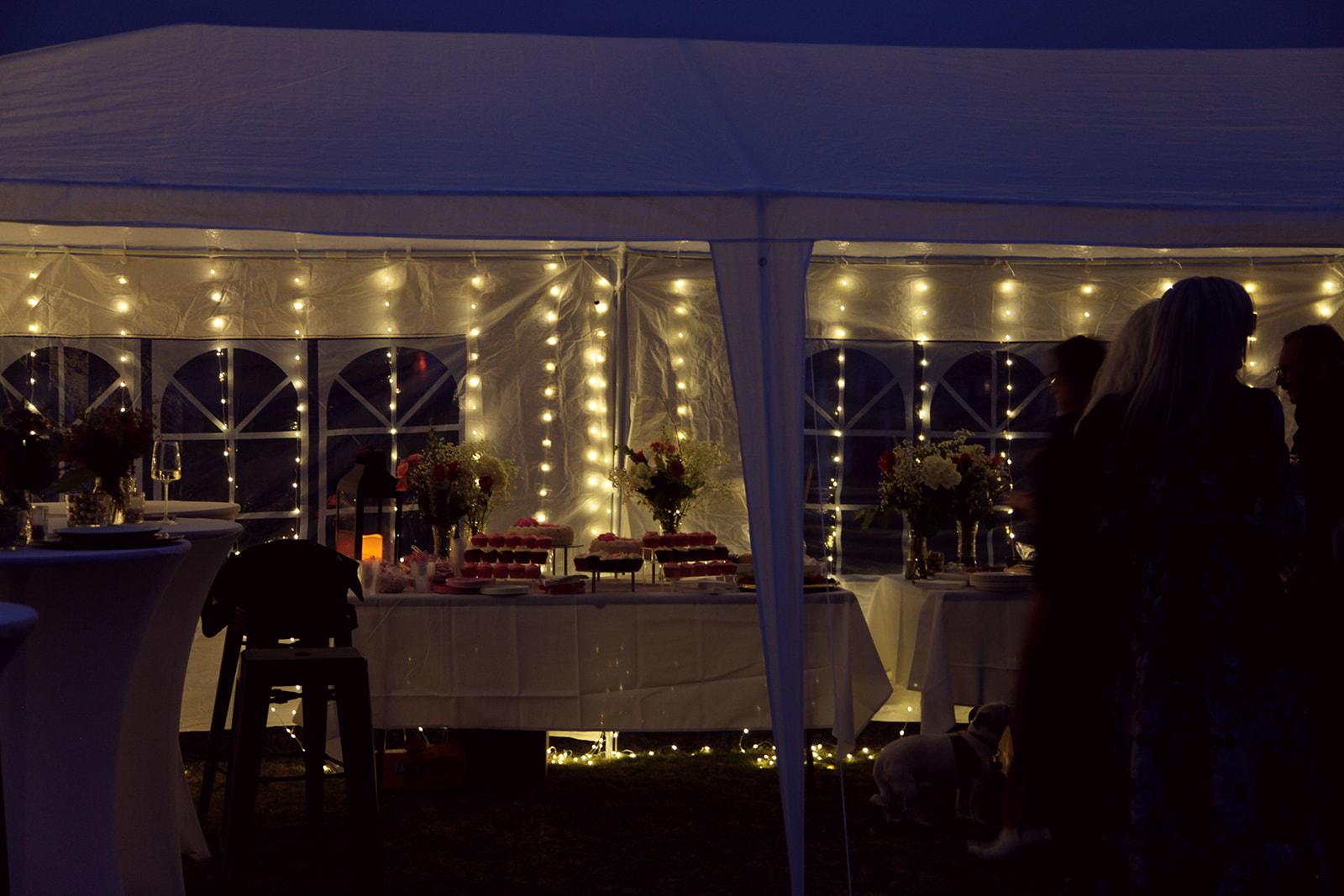 White event tent glowing at dusk with string lights, dessert spread, and guests mingling